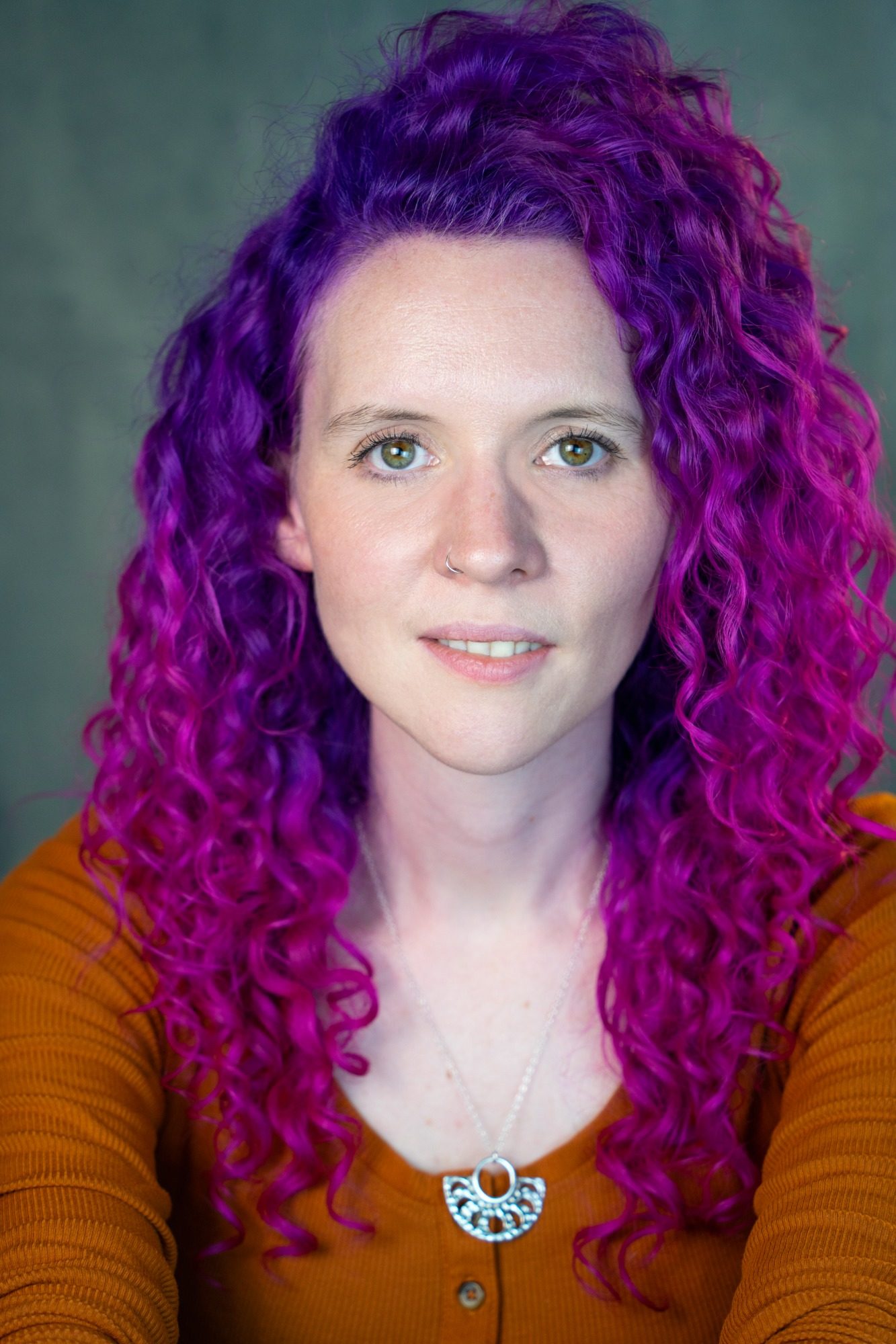 A white woman with pink curly hair wearing an orange top and a silver necklace