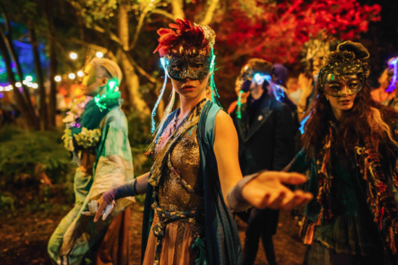 A masked performer standing in the woods reaching out a hand to the camera. Other performers and lights in the background