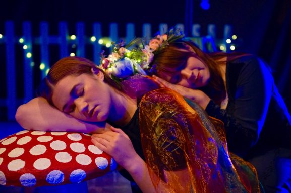 Two women leaning on a mushroom sleeping. One is holding a puppet wrapped in fairy lights