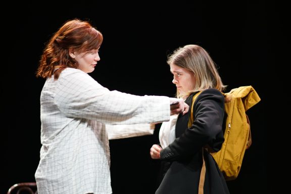 A woman with red hair in a white shirt, reaching out to a woman with blonde hair who is wearing a school uniform and a yellow backpack